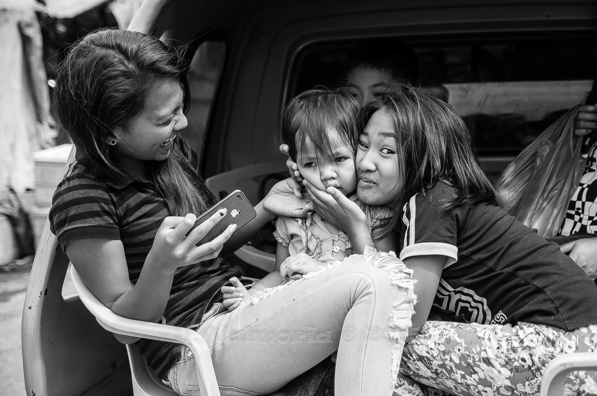 Two young women sit together near the open trunk of a vehicle in black and white, both leaning close to a small child on their lap, sharing a tender moment.