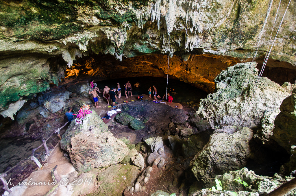 A group of visitors stand on rocky formations inside a large cave cavern in the Philippines, surrounded by stalactite-covered walls and ceiling lit in warm amber tones, with a dark pool of water below.
