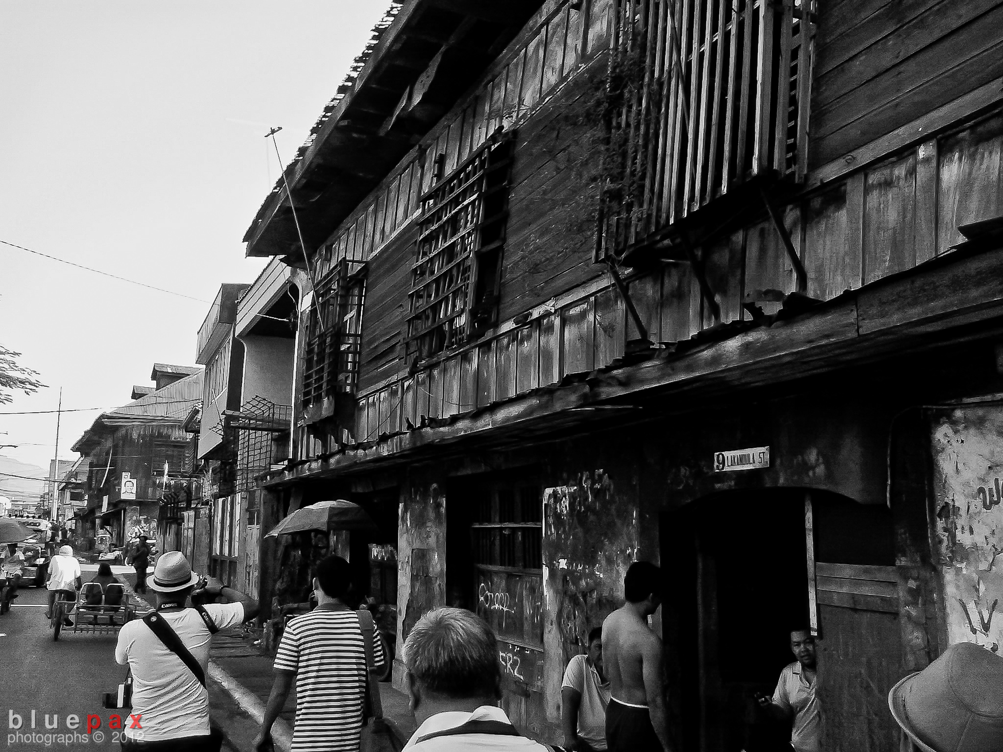 Pedestrians walking along a narrow street beside a deteriorating two-story colonial-era wooden building with peeling walls and graffiti in black and white.