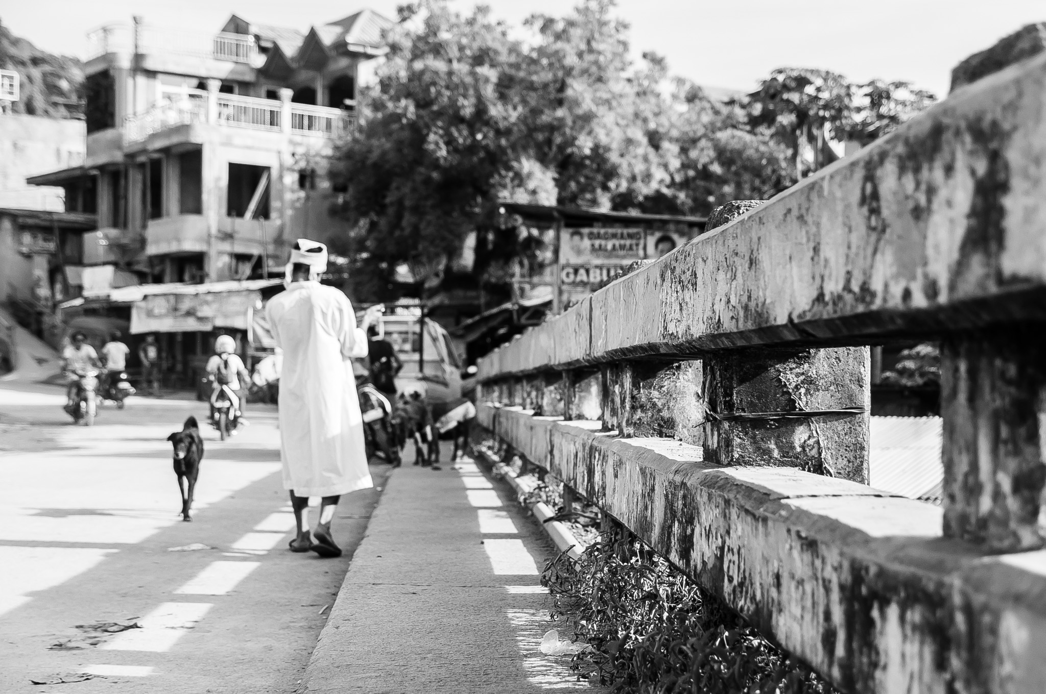 A man dressed in white traditional robes and cap walks along a bridge in a busy Southeast Asian city in black and white, accompanied by a dog, with motorcycles and shophouses in the background.